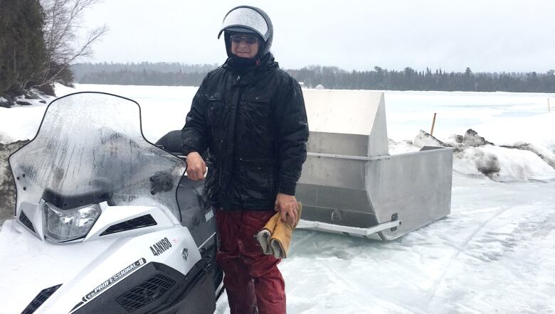 A man stands beside a snowmobile, with a large metal sleigh in behind 