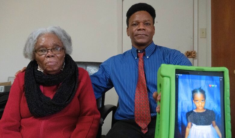 Two people sit in chairs. One holds out a tablet computer with a picture of a child on it.