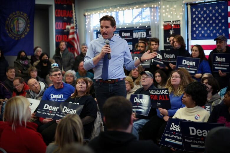 Man in shirtsleeves and tie speaking on stage, surrounded by people and signs that say 'Dean'