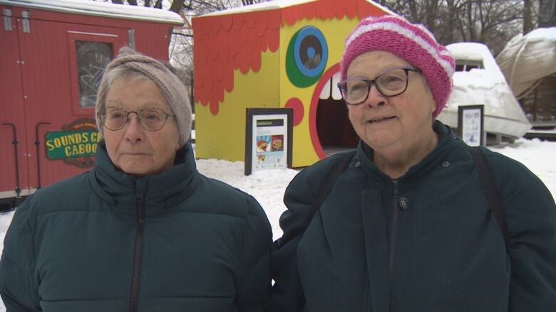 Two older women in winter clothing stand side by side. Behind them are colourful shacks.