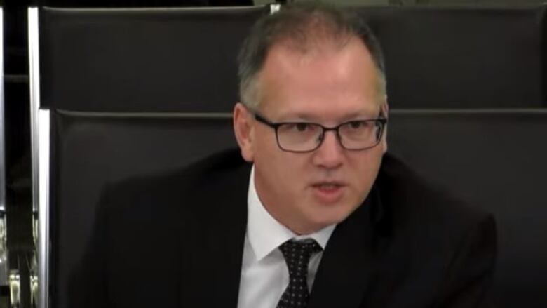 A man with glasses and short hair, waiting a suit, sits at a table.