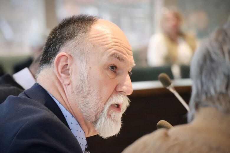 A man with a white beard and moustache, dark hair and a blue suit speaks into a microphone.