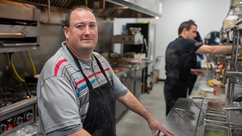 A man in a grey polo shirt and black apron stands in front of a restaurant kitchen counter.