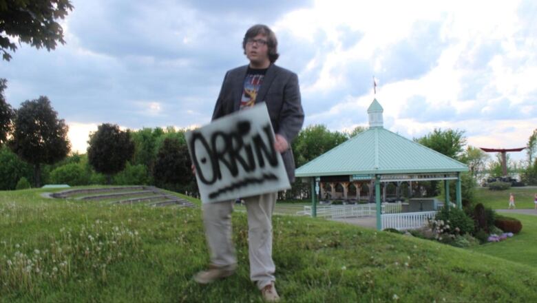 A young man holds a sign that reads 'Orrin' in front of a park bandshell.