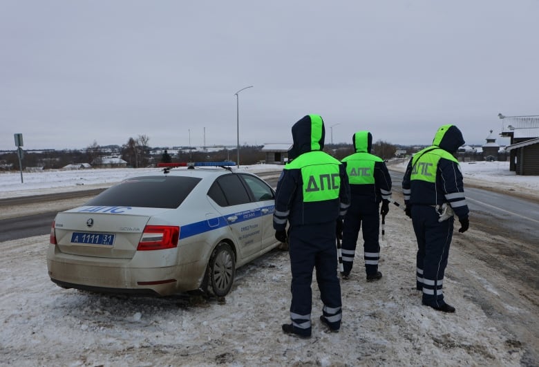 Three people wearing winter coats with hoods over their heads are shown from behind standing on a wintry street near a police vehicle.