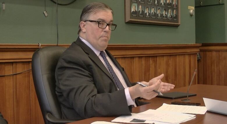 Man in glasses. wearing dark suit, gestures with his hands while seated at a conference table. 