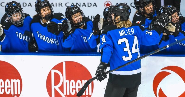 A hockey player in a blue jersey skates along the bench, high fiving her teammates after scoring. 