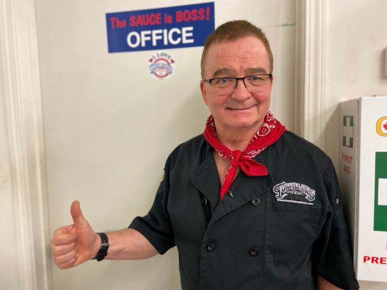 A man holds a thumbs up while wearing a Tumblers Pizza uniform and a hankerchief tied around his neck. 