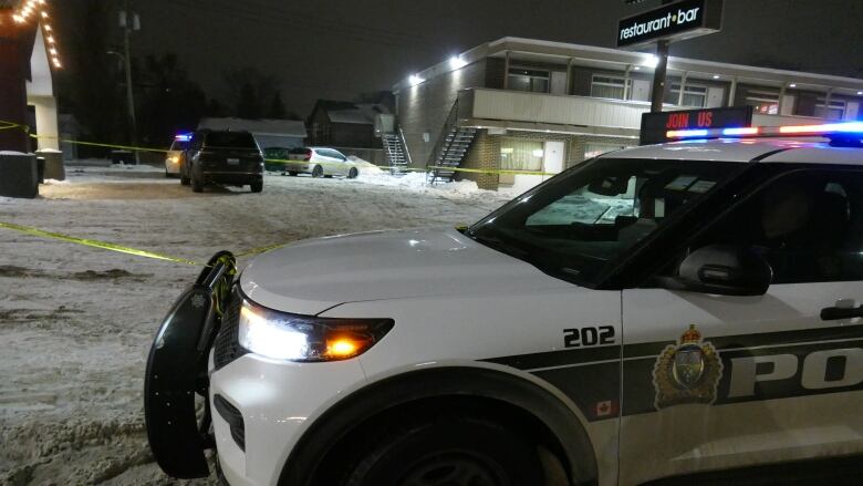 Police car is in foreground on a street, at night in winter. In the background is police tape and a business.