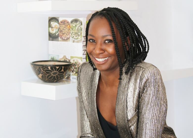 A woman stands in a bright white room, smiling to the camera. She is leaning on a marble counter and there is a book and a vase on a shelf behind her.