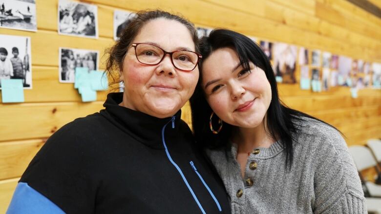 Two women stand smiling in front of a wood-paneled wall.