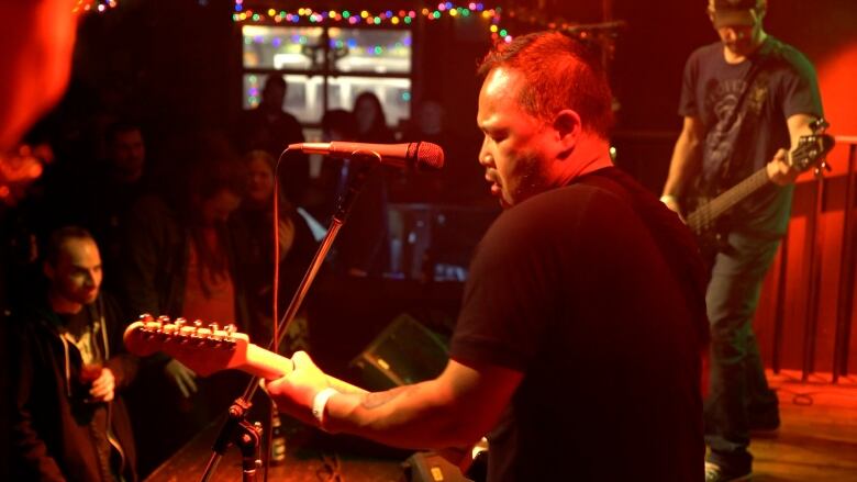 Man standing in front of a microphone and a crowd, playing the guitar on stage.