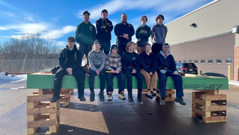 11 people stand in two rows on top of a raised wooden floor. They are outside. The floor is the start of a tiny home that is under construction. 