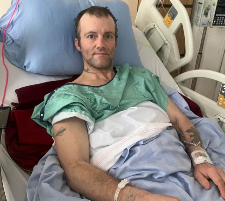 A man with short brown hair, wearing a green medical gown, lying in a hospital bed, covered with blue and white sheets.