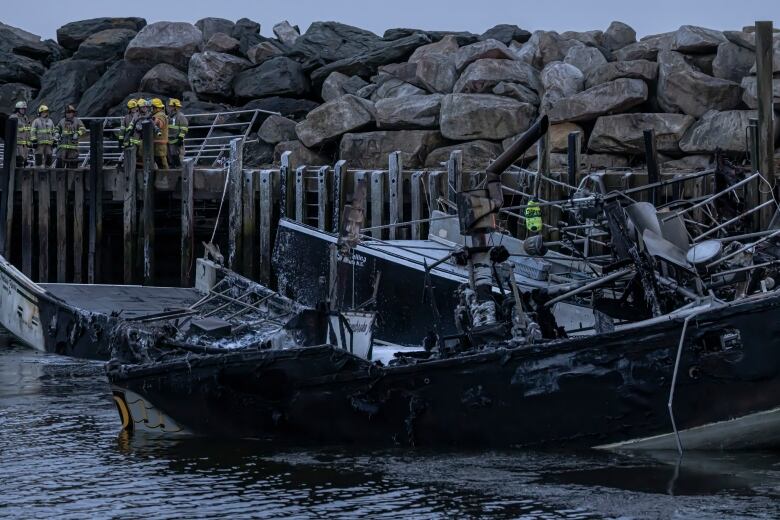 A burned board sits in the water. In the background, there is a group of firefighters