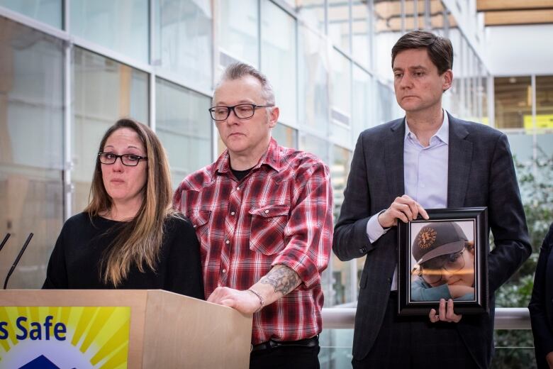 A white man and woman weep at a podium, while a white man behind them holds a picture of a young boy.