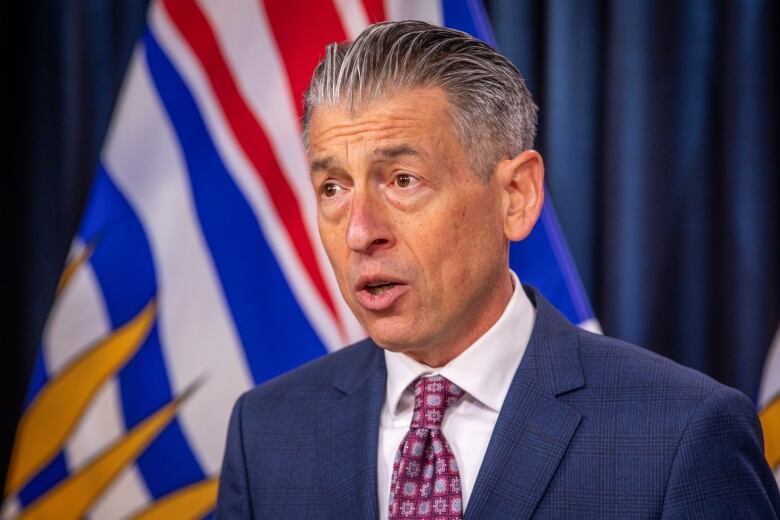 A white man wearing a purple tie stands in front of B.C. flags.