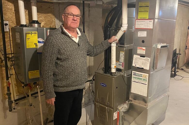 An older man with glasses wearing a grey sweater and dark pants stands in a basement with his hand on a furnace pipe. 