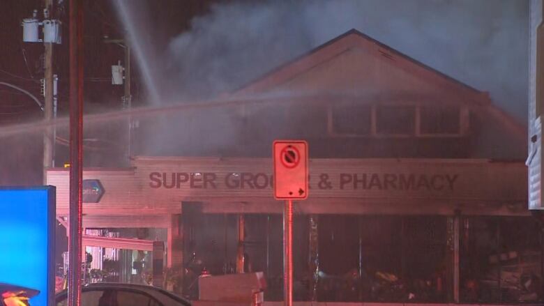 A store reading 'Super Grocer & Pharmacy' has smoke spewing out of it as firefighters spray water on it at night.