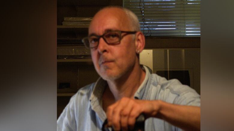 A close-up portrait of a man with bald hair and glasses holding a cane.