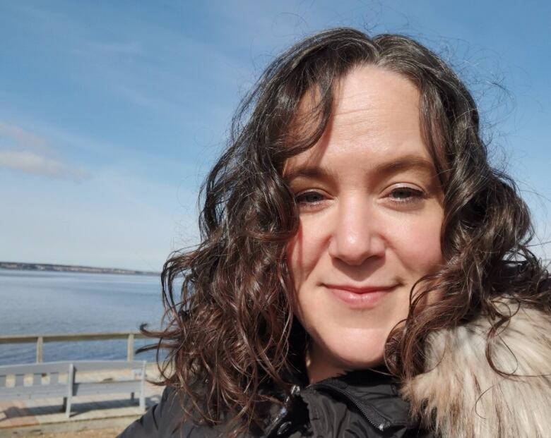 A woman with long hair and wearing a winter coat stands on a dock smiling at the camera.