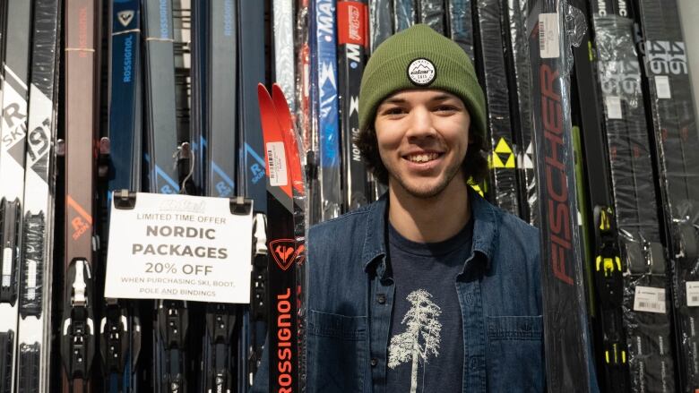 A young man wearing a green toque and a jean jacket smiles for a photo. All around him are skies of different colours.