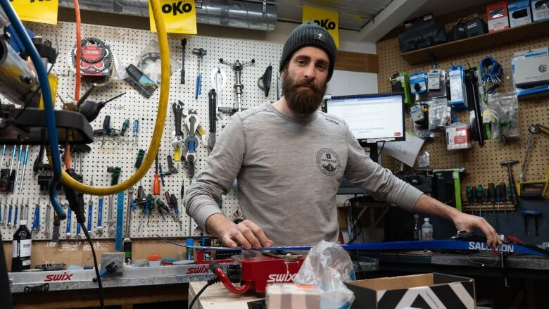 A man wearing a toque and grey shirt waxes a ski inside a workshop. Around him are various tools hung up on a wall.