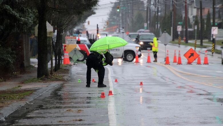 A police offiver with an umbrella bends down to the road where there are several numbered markers. 