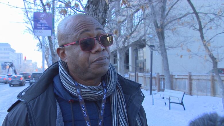 A man in a scarf and sunglasses stands on the sidewalk.