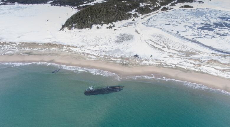 The old hull of a ship sits near the edge of a beach with snowy mountains in the distance.