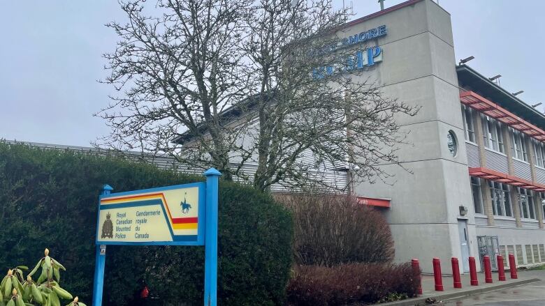 A grey building with a blue sign that says 'West Shore RCMP' partially covered by a barren tree. In the foreground, hedges and a signpost that says 'Royal Canadian Mounted Police' in English and French.