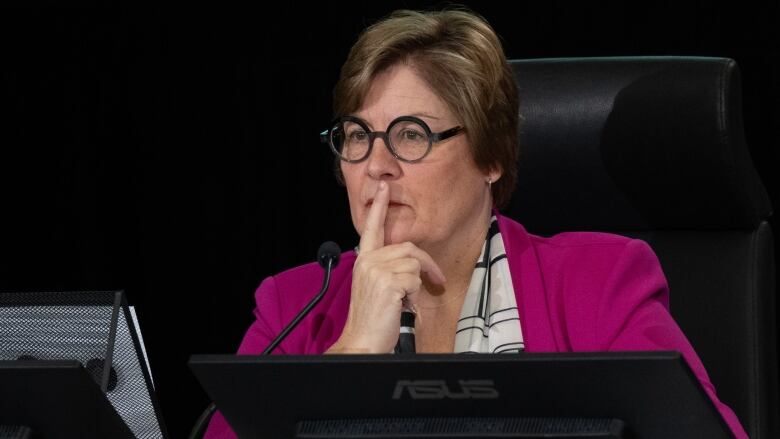 A woman in a pink blazer and glasses poses thoughtfully, while seated in front of a microphone. 