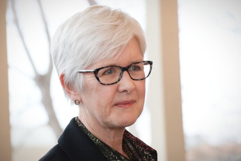 A woman with short white hair and dark framed glasses smiles.