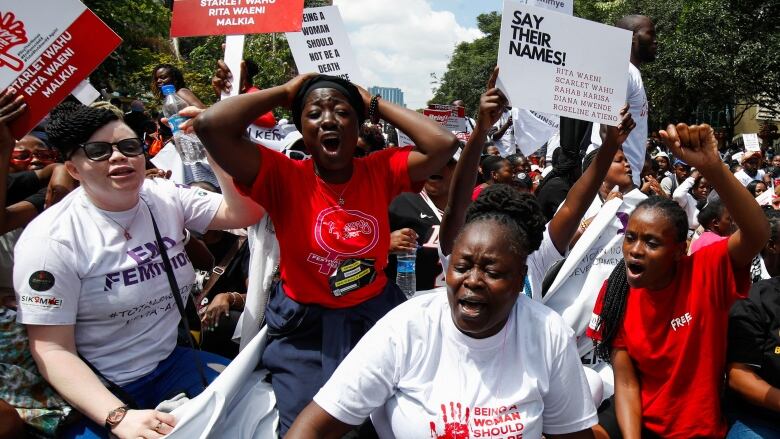 Four women are foregrounded against a crowd of protesters. They are shouting, pumping their firsts or holding their hands against their heads. They wear shirts that say 'End Femicide,' 'Being a Woman Shouldn't Be a Death Sentence' and 'Free.' The protesters behind them hold signs with women's names.