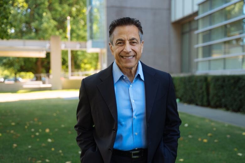 A man with short dark hair, a light blue collared shirt and dark blazer smiles outside with greenery and a grey building in the background.