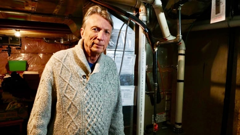 A man with grey hair wearing a grey sweater stands in a basement looking directly at the camera. A furnace, pipes, and aluminum ducts are visible in the background.