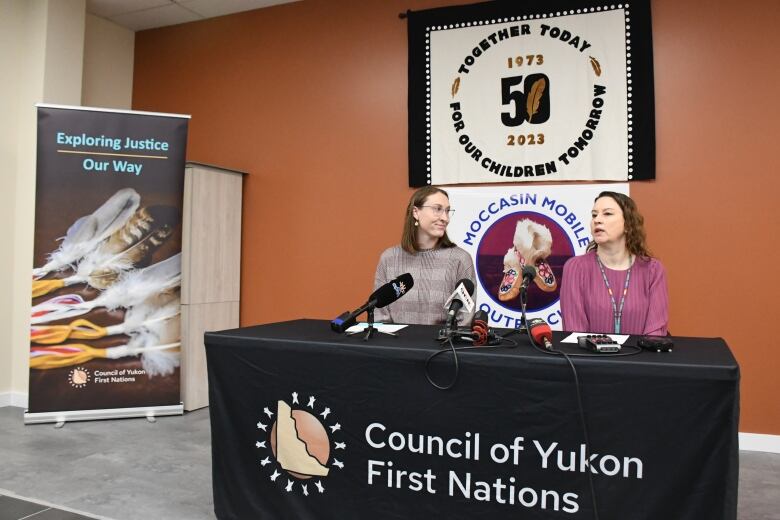 Two women sit at a black table with microphones in front of them. 