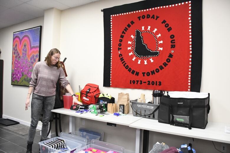 A woman gestures her hand towards tables covered in supplies including naloxone kits, a first-aid kit, harm-reduction supplies and coffee.