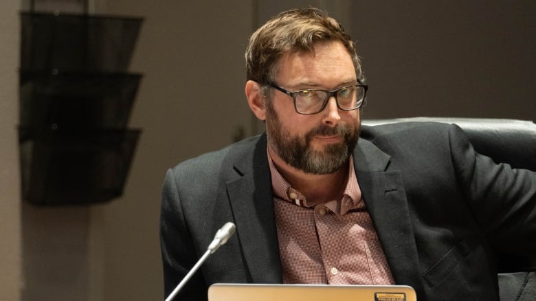 A man in a pink shirt and a grey suit, with glasses, looks sidelong at the camera