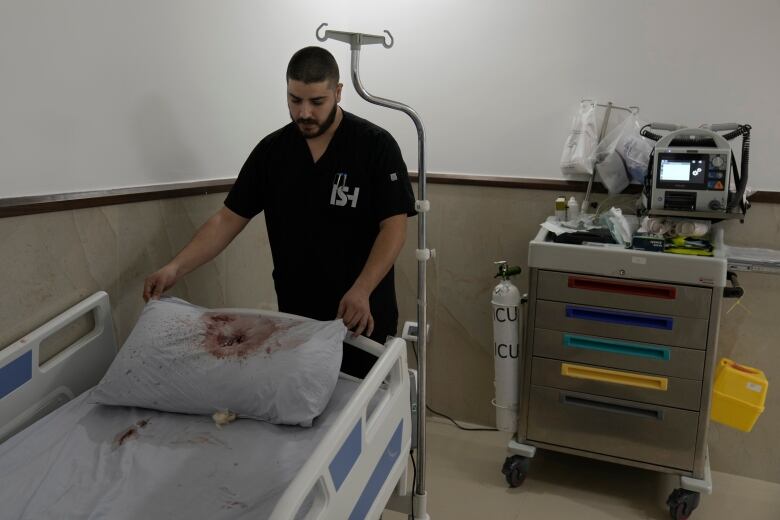 A man in dark hospital scrubs stands above a hospital bed with a blood-stained pillow.