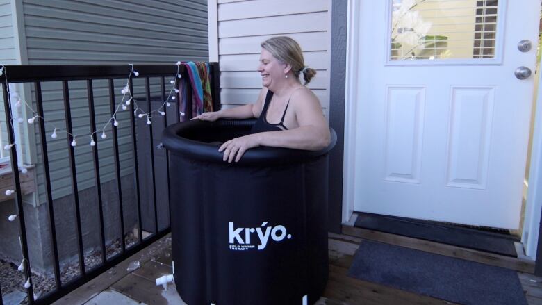 A woman in a bathing suit smiles uncomfortably as she dips herself into a black plastic tub.