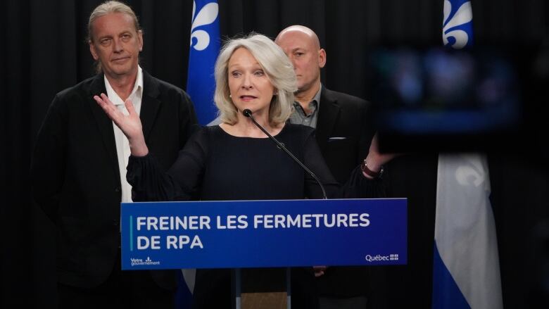 A woman stands at a podium. Behind her are two men. The sign on the podium says slow down the rate of RPA closures. 