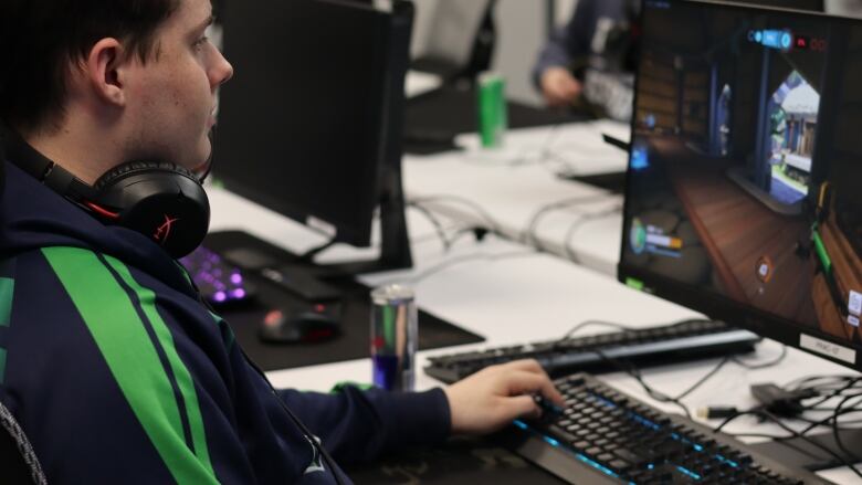 A young man wearing a blue hoodie with a green streak on the sleeve. His one hand is playing with the keys on a light-up keyboard of a desktop computer. 