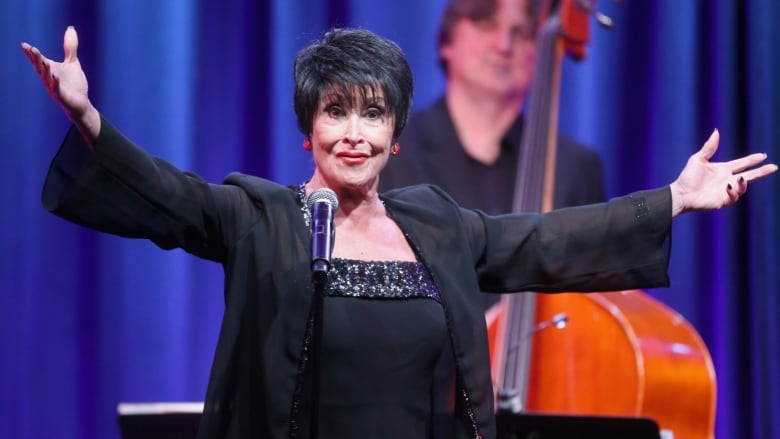 A woman standing in front of a microphone gestures broadly. Behind her is a man at an upright bass. 