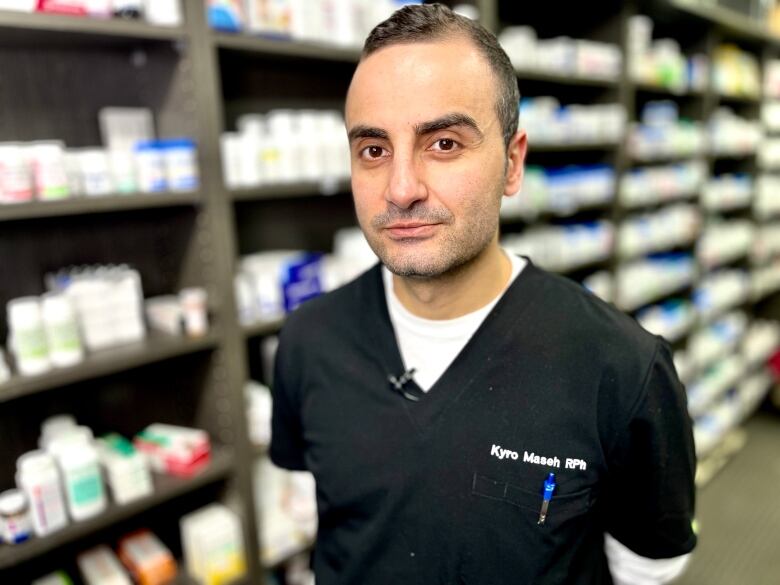 A man wearing pharmacist scrubs is pictured in front of a wall of medications.