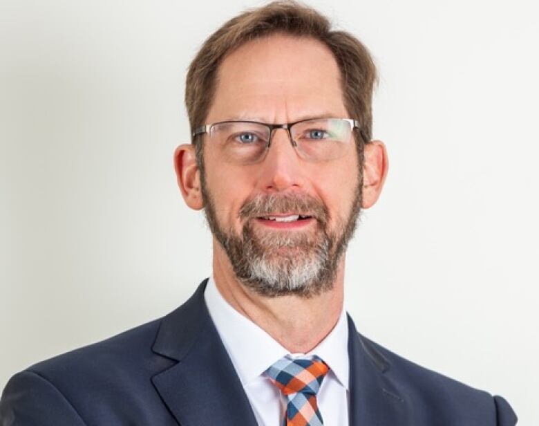 Man with glasses and grey-brown hair and beard wearing navy suit jacket and white shirt with tie.