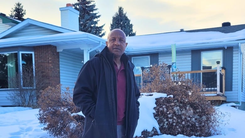 A man stands in the snow in front of a small bungalow.