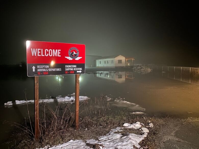 An airport welcome sign in the foreground and a flooded field with a small building in the background. 