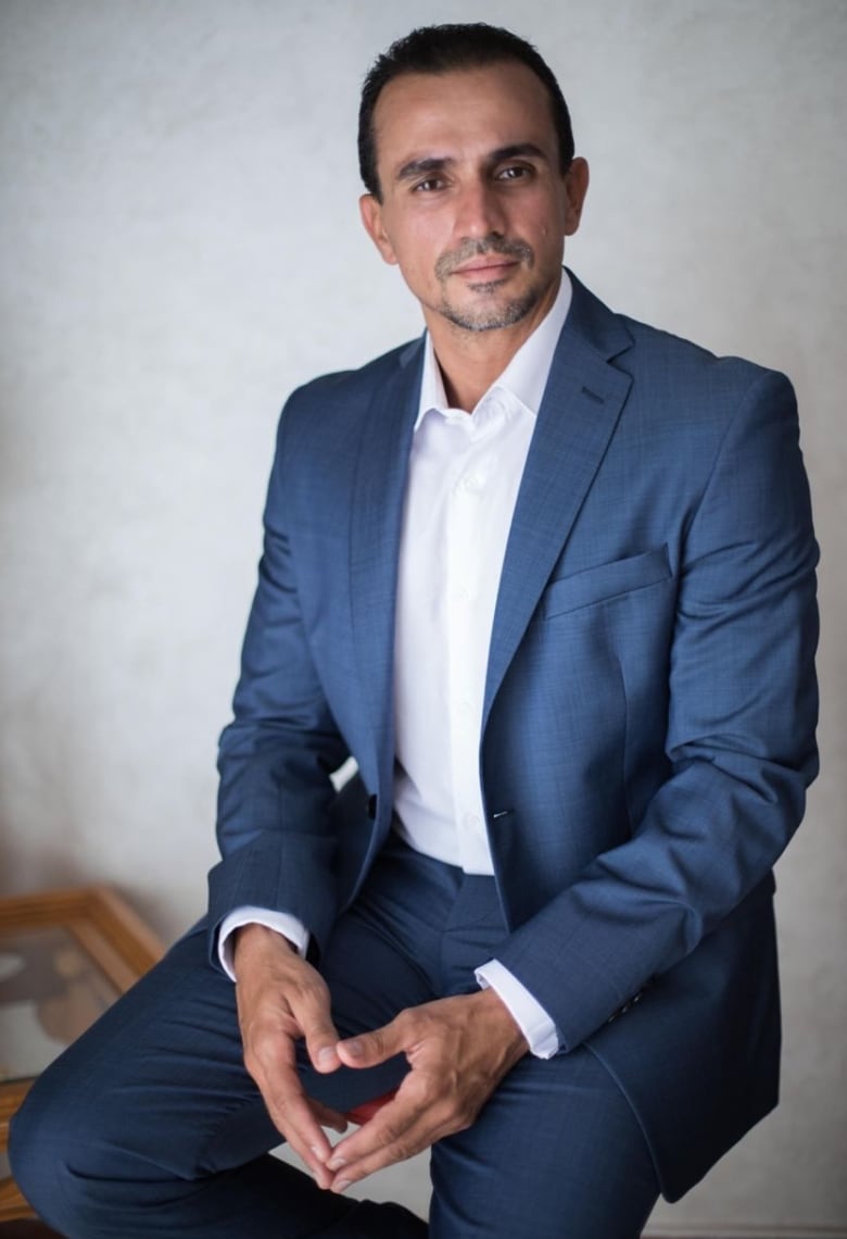 A man wearing a blue suit and white shirt is seated in front of a white wall. 