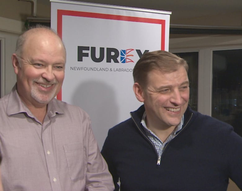 Two smiling men stand in front of a white and red banner that reads 'Furey. Newfoundland and Labrador.'